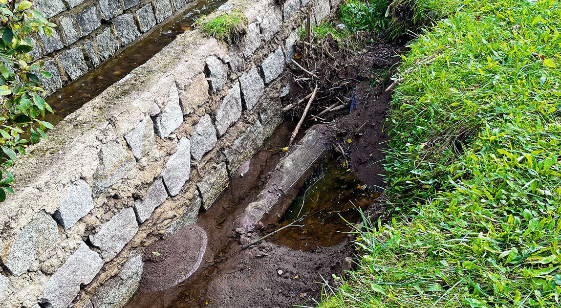 Skutki przesiąków wody od strony cieku do gruntu nasypu w wyniku lokalnych ubytków muru oporowego (wymycie zasypki od strony zewnętrznej)