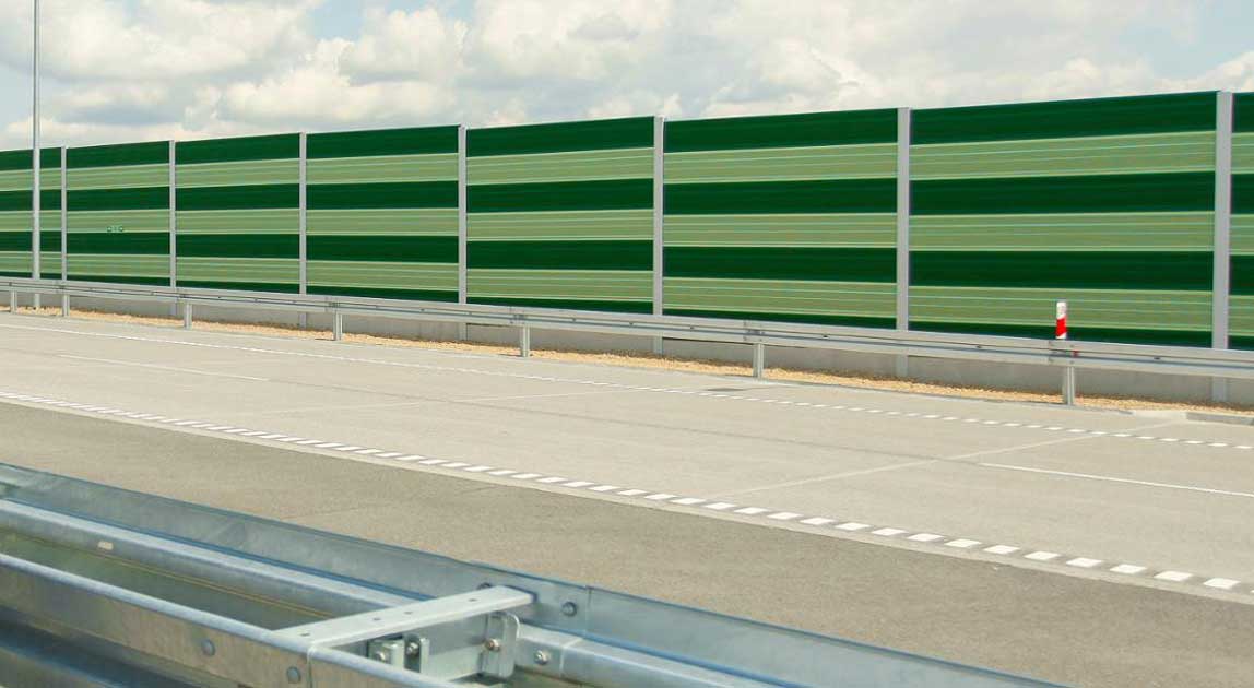 Ekran akustyczny ustawiony wzdłuż autostrady z jezdnią i barierą ochronną.