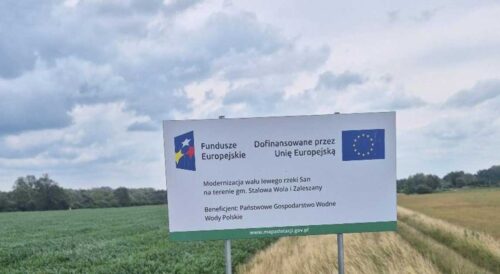 Tablica informacyjna dotycząca modernizacji lewego wału rzeki San na tle terenu inwestycji.