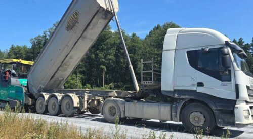 Samochód ciężarowy z wywrotką oraz rozściełacz asfaltu podczas układania nawierzchni drogowej.