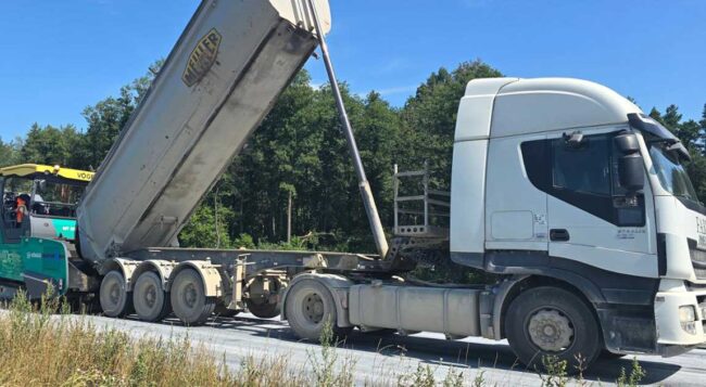 Samochód ciężarowy z wywrotką oraz rozściełacz asfaltu podczas układania nawierzchni drogowej.