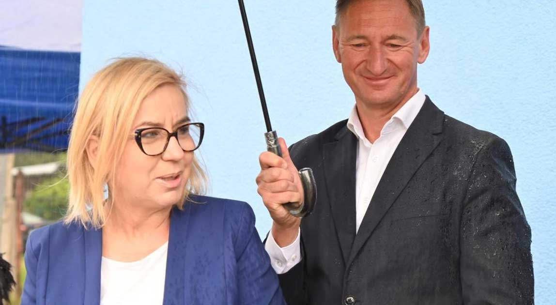 Minister klimatu i środowiska przemawia podczas wydarzenia plenerowego, obok stoi mężczyzna trzymający parasol.